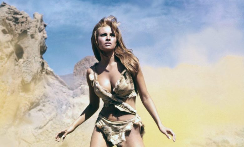 A woman in a primitive-looking, tattered outfit stands confidently against a hazy yellow and blue sky backdrop, with rocky terrain visible. Her long blonde hair blows in the wind, and she looks int