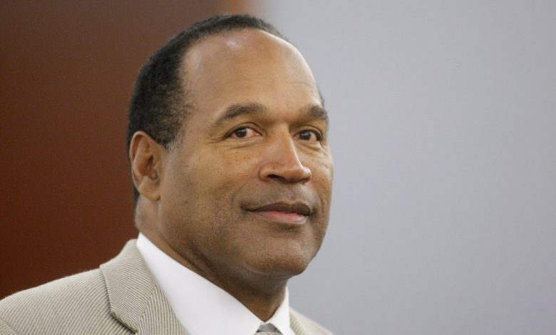 A middle-aged African-American man in a courtroom, resembling O.J. Simpson, wearing a tan suit and tie, looks slightly to his right with a serious expression. He has short black hair and is