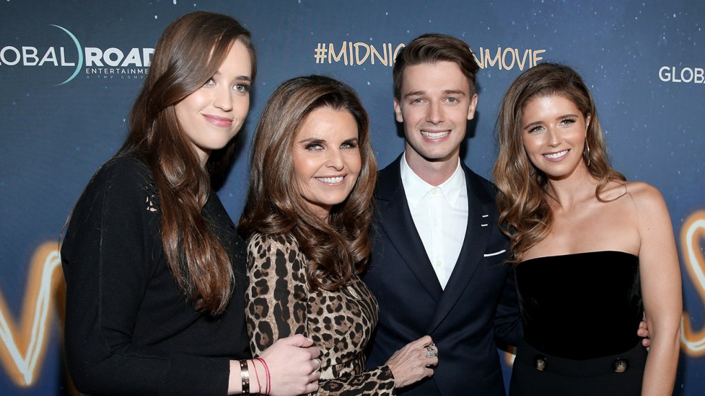 Four adults smiling at a movie premiere, standing closely together in formal attire against a backdrop with logos and #midnightmovie. The women wear elegant dresses, and the man is in a suit. A