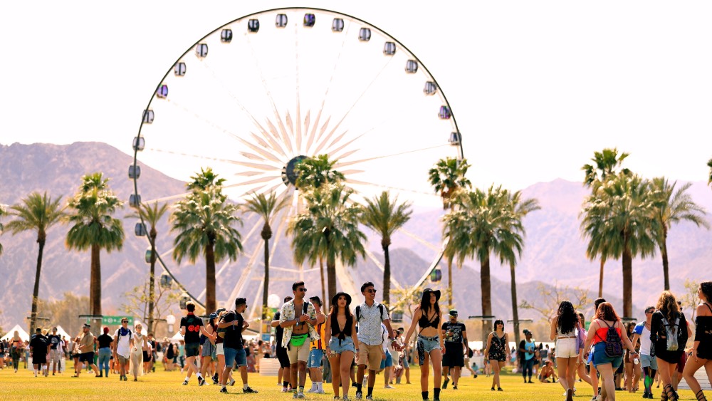 Young adults walk across a grassy area at Coachella 2024 music festival, with a large ferris wheel and palm trees against a mountainous backdrop under a clear sky.