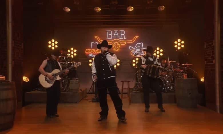 Grupo Frontera performs on stage with warm lighting. Three musicians play guitar and drums, with a vocalist in the center wearing a black hat and jacket. The backdrop features a neon "bar" sign