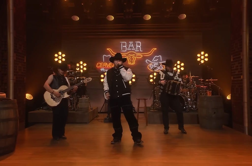 Grupo Frontera performs on stage with warm lighting. Three musicians play guitar and drums, with a vocalist in the center wearing a black hat and jacket. The backdrop features a neon "bar" sign