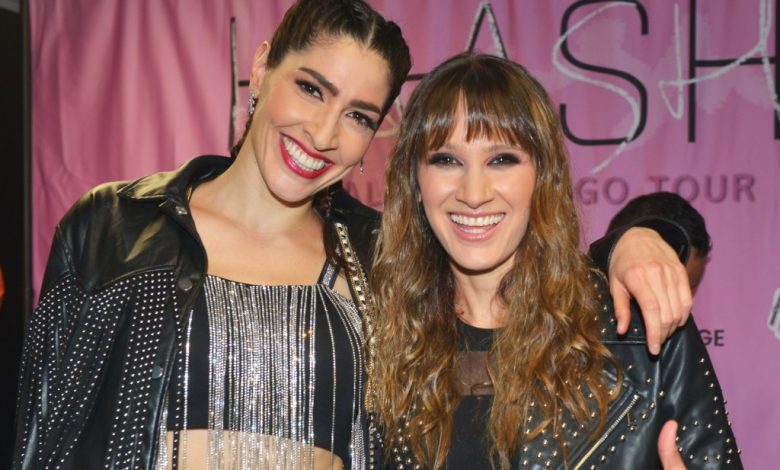 Two women, possibly Latin music siblings from Ha*Ash, smiling at a promotional event, one with a high ponytail and metallic top, and the other with fringed bangs; both in black