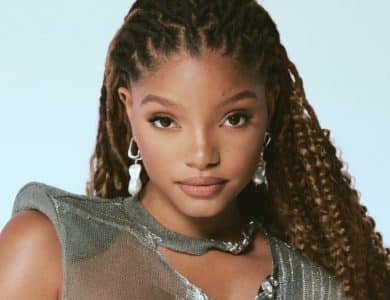 A young woman with braided hair and silver earrings poses against a light blue background. She wears a metallic sleeveless top and has a subtle smile, reminiscent of Halle Bailey's charm.