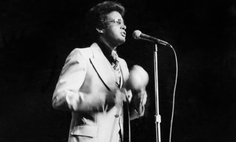 A black and white photo of a man in a suit and glasses passionately singing into a microphone on a dark stage, holding a tambourine, embodying a soulful expression and dynamic performance, capturing