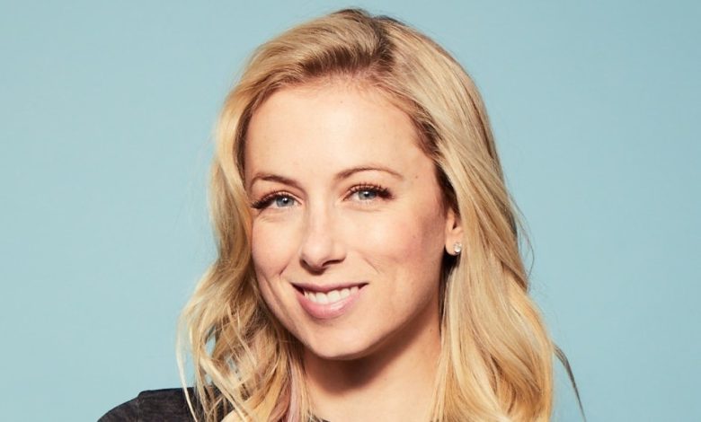 A close-up portrait of a smiling woman with long blonde hair, wearing a black top against a light blue background. She has fair skin and earrings, presenting a friendly appearance as she promotes Prime Video's