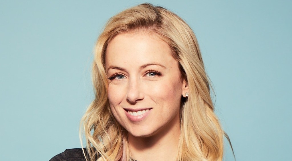 A close-up portrait of a smiling woman with long blonde hair, wearing a black top against a light blue background. She has fair skin and earrings, presenting a friendly appearance as she promotes Prime Video's