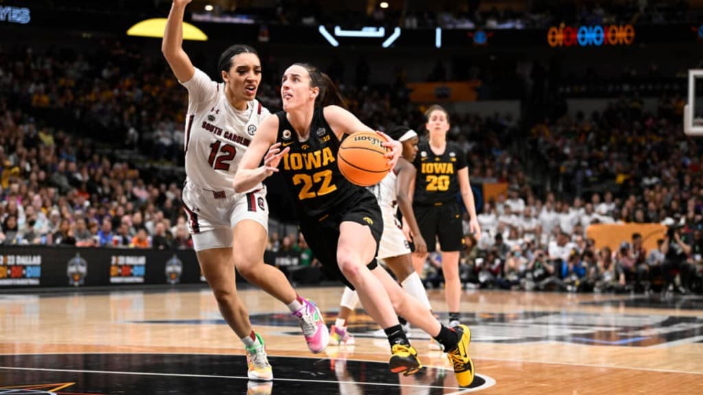 Two female basketball players in action during the 2024 NCAA Women's Final. A South Carolina player wearing a white jersey, number 12, competes for the ball against an Iowa player in a