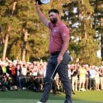 A male golfer in a maroon shirt and black pants waves his cap to a cheering crowd, bathed in the warm glow of a setting sun behind towering pine trees at the Masters Golf Tournament.
