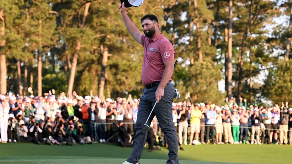 A male golfer in a maroon shirt and black pants waves his cap to a cheering crowd, bathed in the warm glow of a setting sun behind towering pine trees at the Masters Golf Tournament.