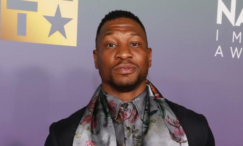 A man stands confidently at the NAACP Image Awards, dressed in a dark gray suit with a detailed floral scarf. His expression is cheerful, offering a slight smile as he poses for photographers.