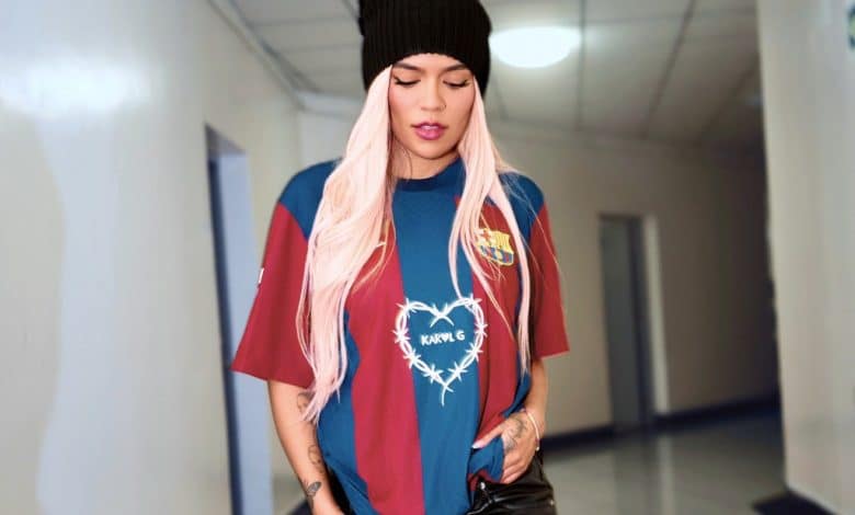A fashionable young woman walks confidently wearing a beanie and a colorful t-shirt featuring Karol G's logo, paired with black leather pants, indoors in a well-lit corridor.