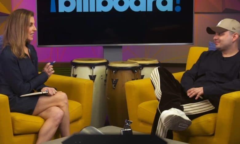 A man and a woman sit facing each other in a studio with "billboard" logos. the woman holds a clipboard, while the man, dressed casually, rests his hands on his thighs, with conga drums nearby.