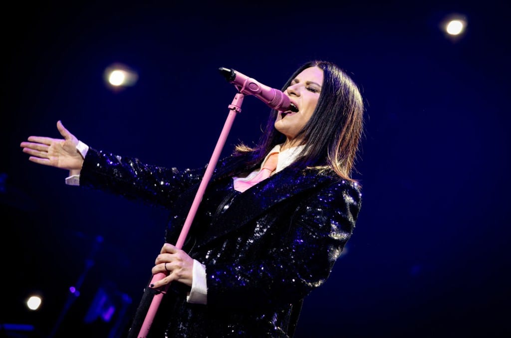 A female singer on stage, passionately performing into a pink microphone. She wears a sparkling black blazer, her long dark hair flowing, under dramatic stage lights as she duets with Fonsi in