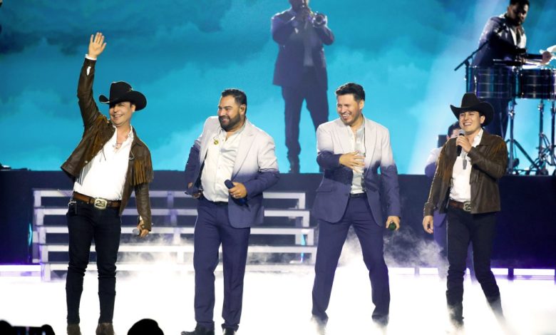 Four male performers on stage at a concert. two wear cowboy hats and dark jackets, while the others are in suits. one waves to the crowd, another plays the trumpet, and a third is on drums in the background. mist and blue lighting enhance the scene.