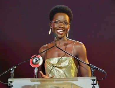 A joyful woman with short hair speaks at a podium, wearing a glamorous golden strapless dress, holding an award. She stands against a blurred red and pink stage backdrop, illuminated by tears