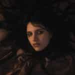 A woman with dark hair and pale skin lies on a bed, her hair spread out around her head. She appears troubled or contemplative, gazing upwards with a somber expression under dim, warm