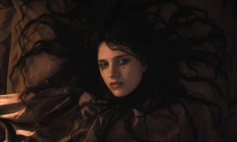 A woman with dark hair and pale skin lies on a bed, her hair spread out around her head. She appears troubled or contemplative, gazing upwards with a somber expression under dim, warm