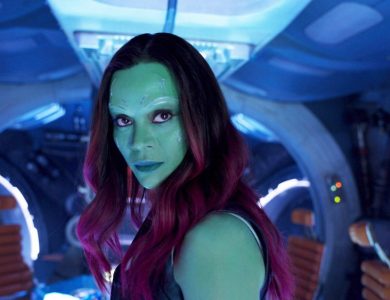 A woman with green skin and vibrant red hair looks intently forward in a dimly lit spaceship cockpit with blue lighting. She has detailed facial markings and appears focused and determined, embodying the intensity Zoe