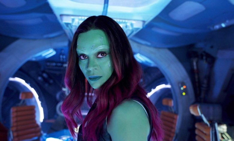 A woman with green skin and vibrant red hair looks intently forward in a dimly lit spaceship cockpit with blue lighting. She has detailed facial markings and appears focused and determined, embodying the intensity Zoe