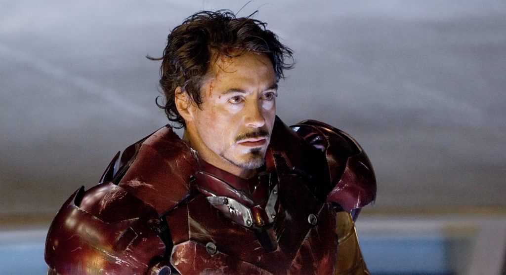 Tony Stark, portrayed by Robert Downey Jr., wearing a damaged Iron Man suit, looks intensely forward with a slight frown, highlighting a scene of tension and eager determination.