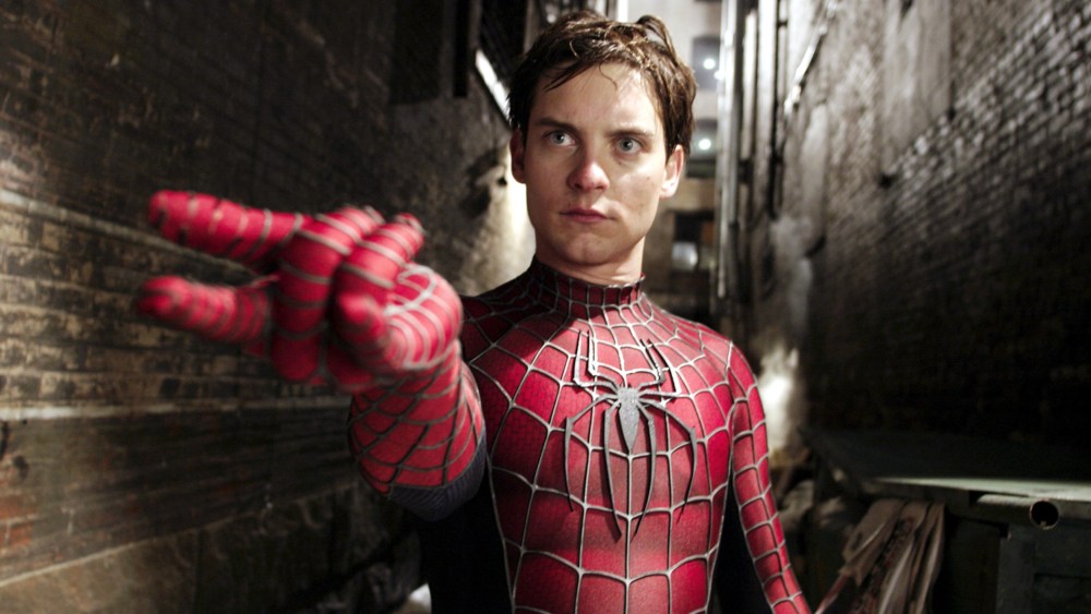 A man dressed as Spider-Man stands in a dark alley, extending his hand forward to mimic shooting a web, with a focused expression on his face. He wears the iconic red and blue Raimi