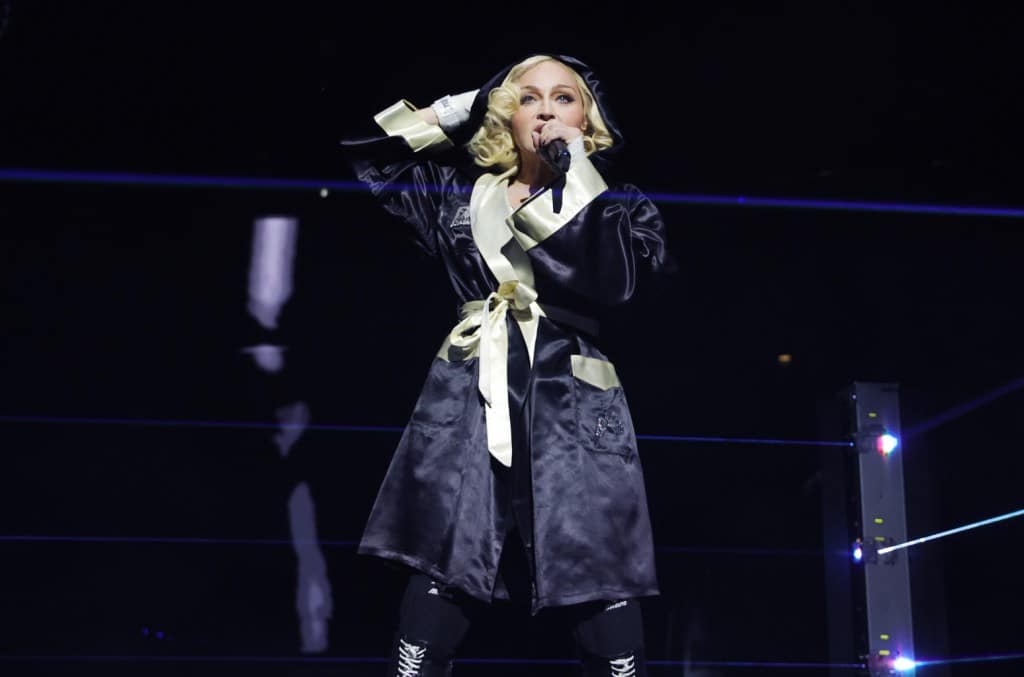 A female performer sings into a microphone on a dimly lit stage, dressed in an oversized black robe with white trim and bow details, accentuated by dramatic stage lighting.