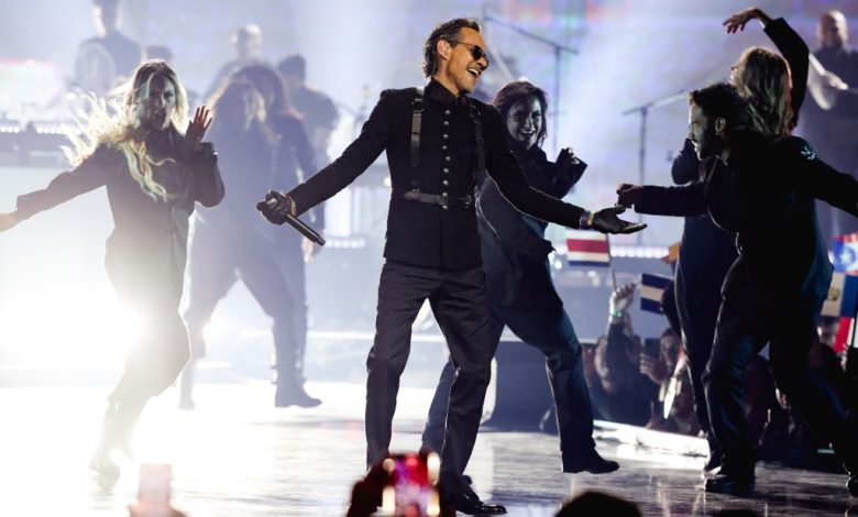 Marc anthony performs on stage, smiling, with arms outstretched, surrounded by energetic dancers in motion under bright stage lights, with musicians visible in the background.