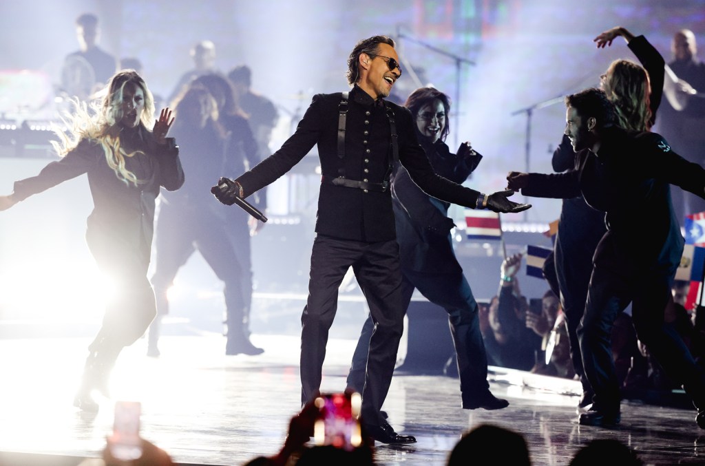 Marc anthony performs on stage, smiling, with arms outstretched, surrounded by energetic dancers in motion under bright stage lights, with musicians visible in the background.