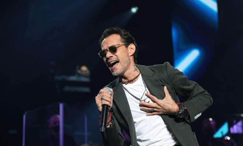 A male singer passionately performing onstage, holding a microphone in his right hand. he has a cross tattoo on his neck, wears glasses, and dons a gray blazer over a black shirt. the background features blue stage lights.