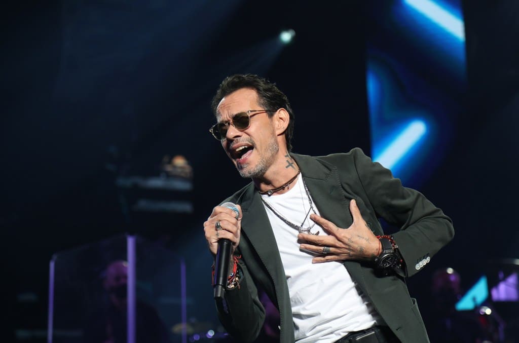 A male singer passionately performing onstage, holding a microphone in his right hand. he has a cross tattoo on his neck, wears glasses, and dons a gray blazer over a black shirt. the background features blue stage lights.