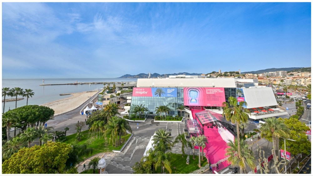 Aerial view of the Cannes Film Festival venue, with large posters on the facade, a sandy beach on the left, marina extending into the sea, and a town with green hills in the background
