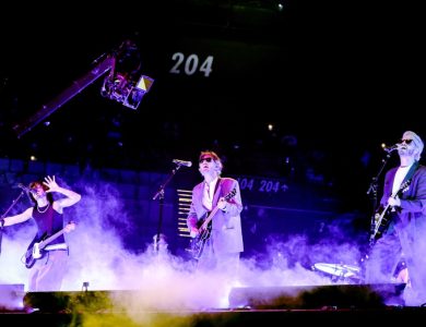 Three musicians perform energetically on a concert stage surrounded by smoke. the vocalist in the center wears a suit and sunglasses, flanked by a guitarist on the left and a bassist on the right, under vibrant stage lighting.