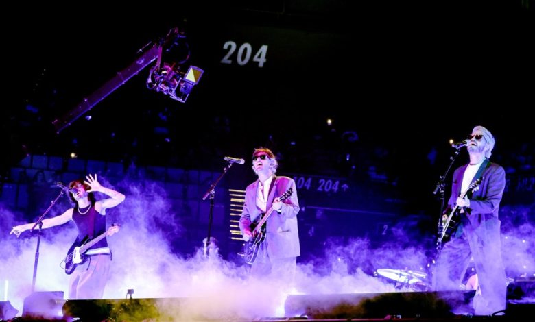 Three musicians perform energetically on a concert stage surrounded by smoke. the vocalist in the center wears a suit and sunglasses, flanked by a guitarist on the left and a bassist on the right, under vibrant stage lighting.