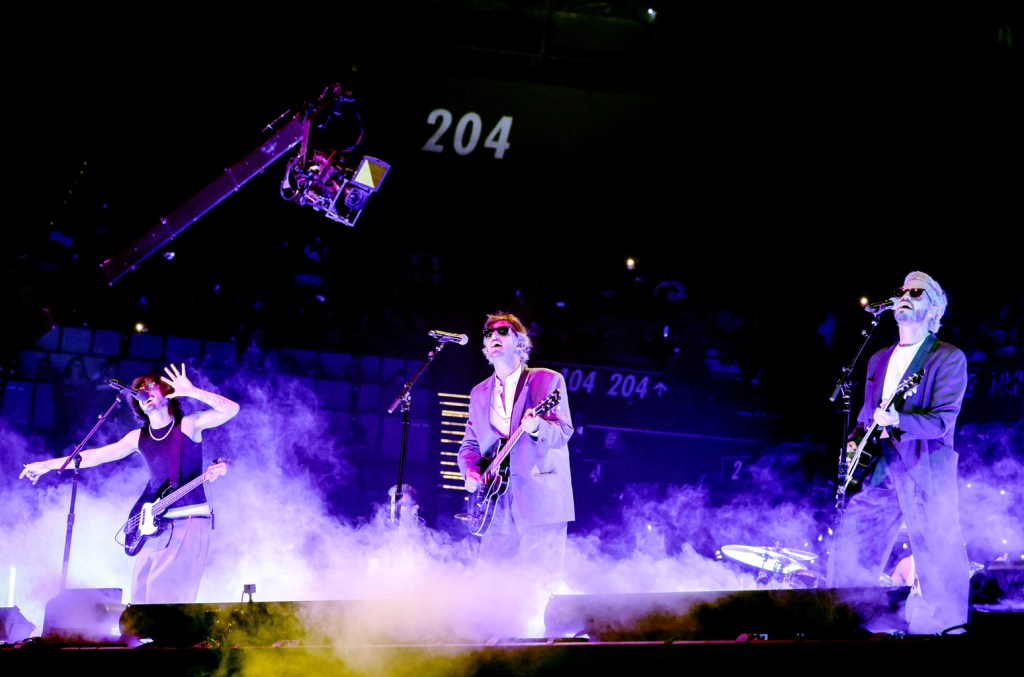 Three musicians perform energetically on a concert stage surrounded by smoke. the vocalist in the center wears a suit and sunglasses, flanked by a guitarist on the left and a bassist on the right, under vibrant stage lighting.