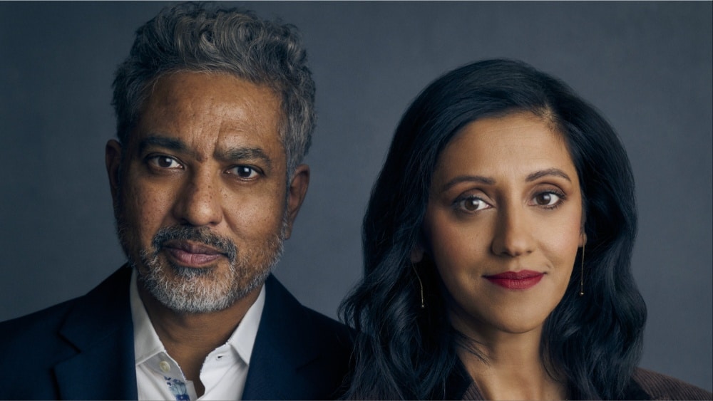 A middle-aged man with graying hair and a woman with long dark hair, both of South Asian descent, stand side by side looking into the camera against a slate backdrop.