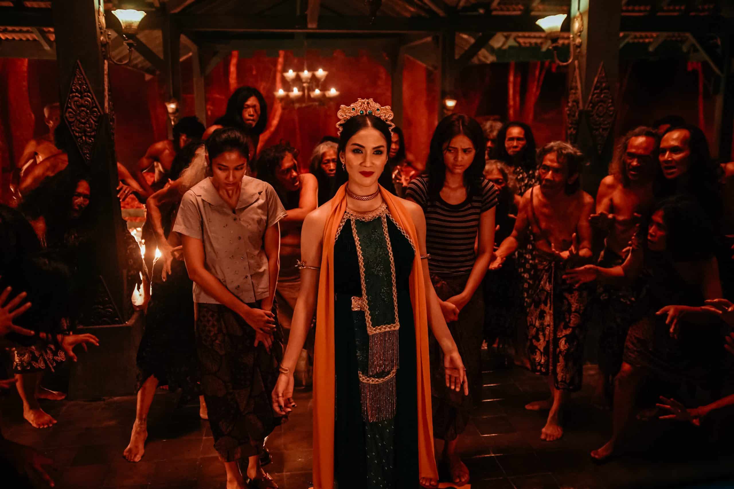 A woman in traditional attire walks confidently at the forefront of a candle-lit ceremony, surrounded by a group of attentive participants in a dimly-lit, ornate village setting.