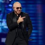 A bald man wearing sunglasses and a black suit gestures with one hand while smiling broadly during a performance on the brightly lit "After Dark Tour" stage.