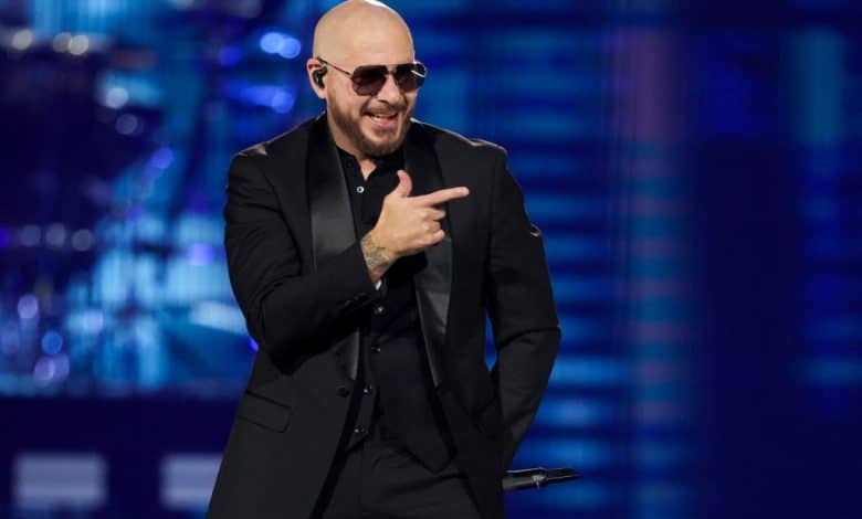 A bald man wearing sunglasses and a black suit gestures with one hand while smiling broadly during a performance on the brightly lit "After Dark Tour" stage.