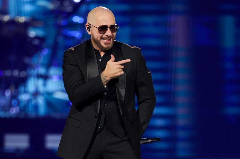 A bald man wearing sunglasses and a black suit gestures with one hand while smiling broadly during a performance on the brightly lit "After Dark Tour" stage.