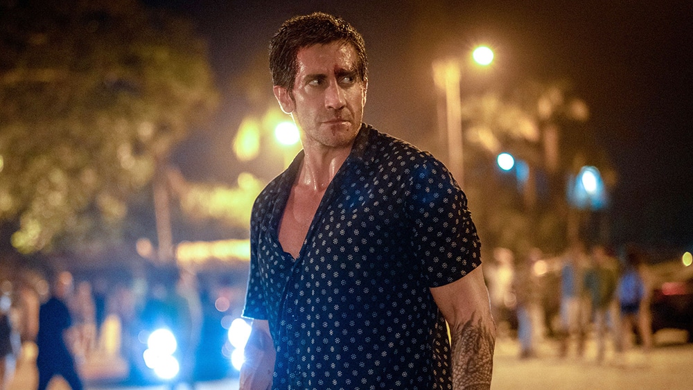 A man with a rugged expression, wearing a dark polo with a white dot pattern, walks at night. The road behind him is dimly lit with blurred lights from vehicles and street lamps.