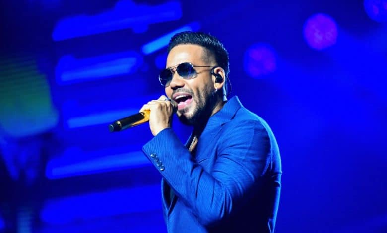 A male singer performs live onstage, capturing the audience's attention with a microphone in hand. He wears a stylish blue suit and sunglasses while singing passionately under vibrant stage lighting. Romeo Santos denies heart attack rumors