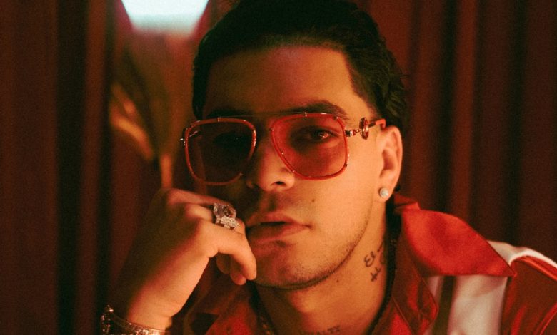 A stylish young man wearing tinted sunglasses and a red bandana under a red light poses against a blurry red backdrop. His head rests lightly on his hand, adorned with silver rings as he listens int