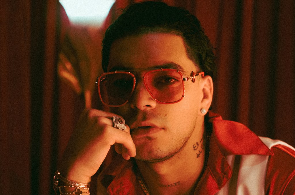 A stylish young man wearing tinted sunglasses and a red bandana under a red light poses against a blurry red backdrop. His head rests lightly on his hand, adorned with silver rings as he listens int
