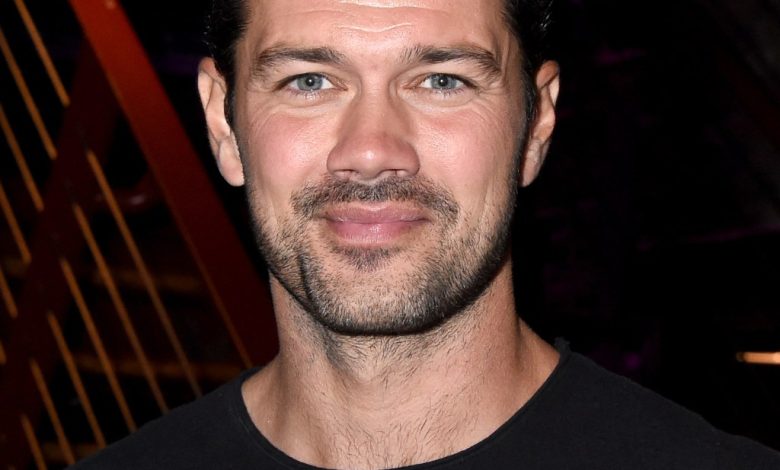 A portrait of a smiling Ryan Paevey with short, wavy black hair and light stubble. He wears a black t-shirt and stands in front of a blurred background with orange geometric patterns.
