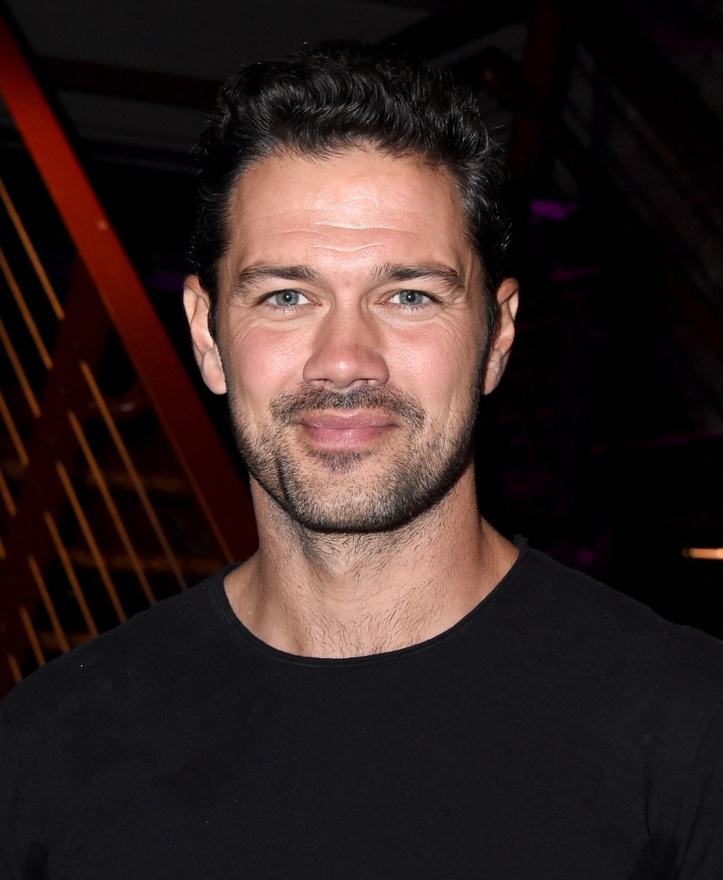 A portrait of a smiling Ryan Paevey with short, wavy black hair and light stubble. He wears a black t-shirt and stands in front of a blurred background with orange geometric patterns.