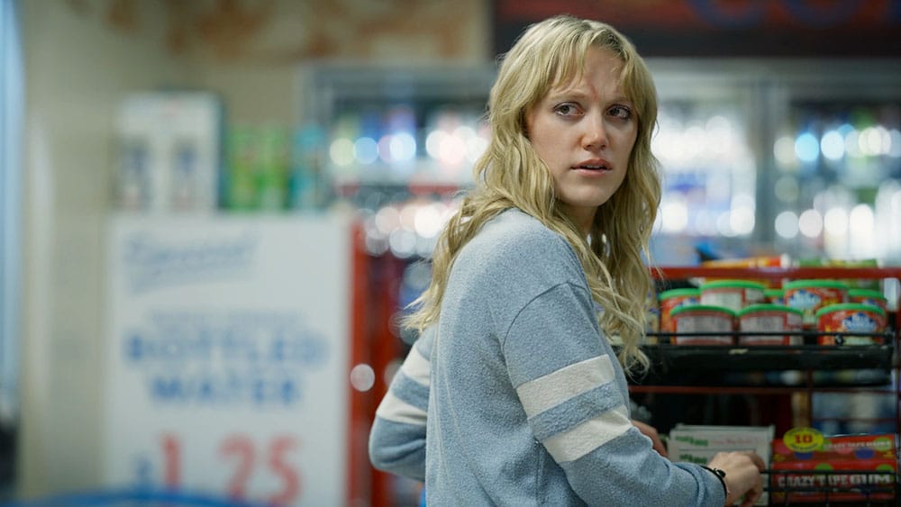 A woman with blonde hair looks over her shoulder with a surprised expression in a grocery store, standing near shelves stocked with snacks, including Veena Sud's Transition. She wears a gray striped sweater.