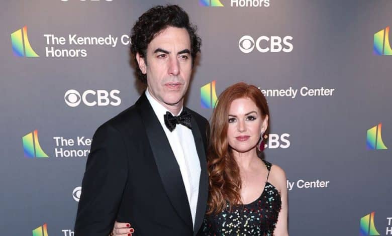 A woman with long red hair and a sequined dress poses next to Sacha Baron Cohen, who is wearing a black tuxedo with a bow tie, standing together at a formal event. Both