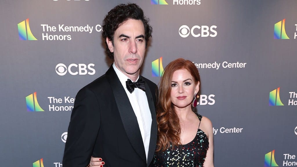 A woman with long red hair and a sequined dress poses next to Sacha Baron Cohen, who is wearing a black tuxedo with a bow tie, standing together at a formal event. Both