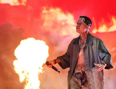 Santa Fe Klan, a tattooed rapper, performs on stage with a microphone, wearing a denim jacket and headscarf, against a backdrop of dramatic red flames and smoke at Coachella.